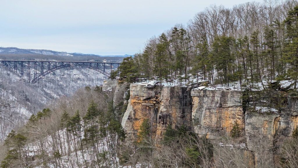 The New River Gorge Bridge in the distance with white snow covered cliffs of the Endless Wall Trail in the foreground. Green pine trees grow on top the cliffs. 