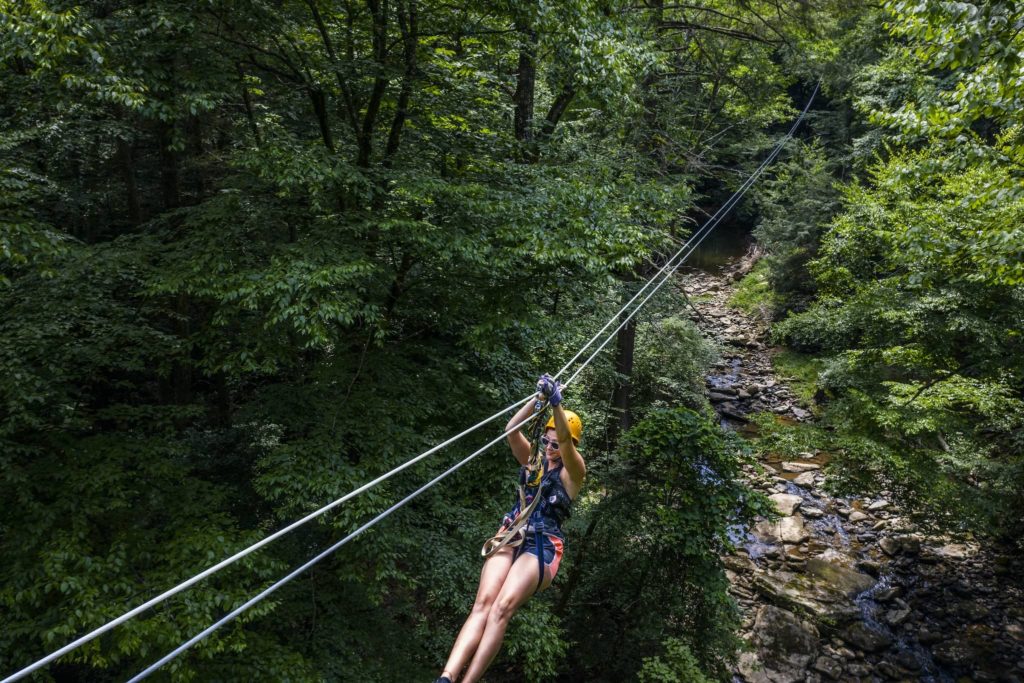 Woman in yellow helmet attached to a zipline high above a creek with trees below her. 