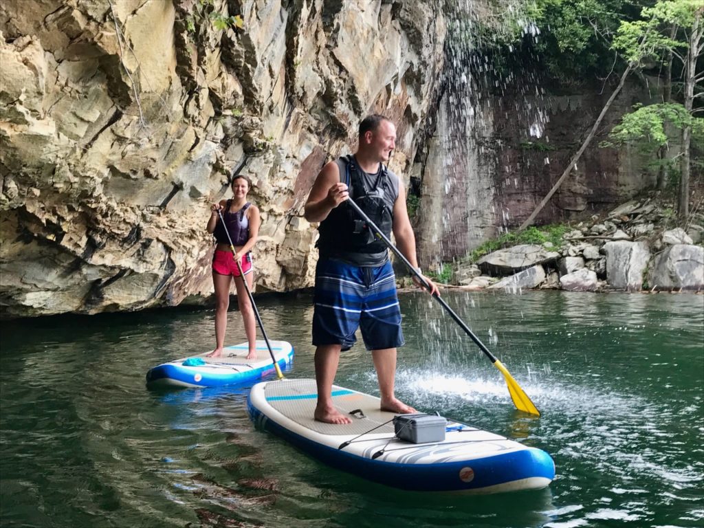 Husband and wife on paddle boards under a waterfall in the New River with rock cliffs in the background. 