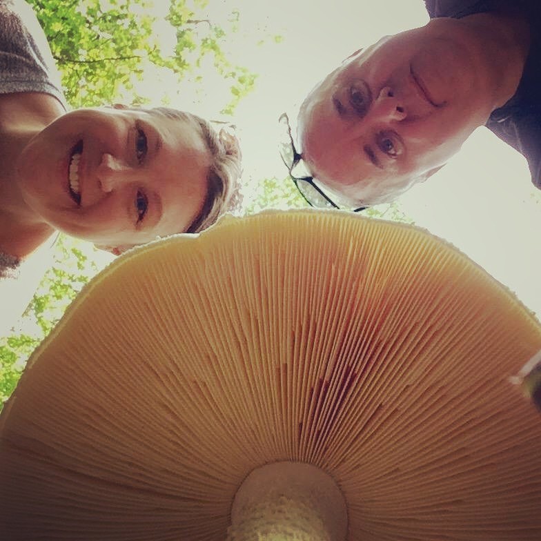 Amy and Shawn smiling above a cream colored mushroom with big well-defined gills. 