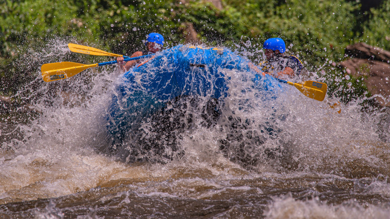 People in bright blue helmets with yellow oars in a raft going through wihitewater rapids on the New River.