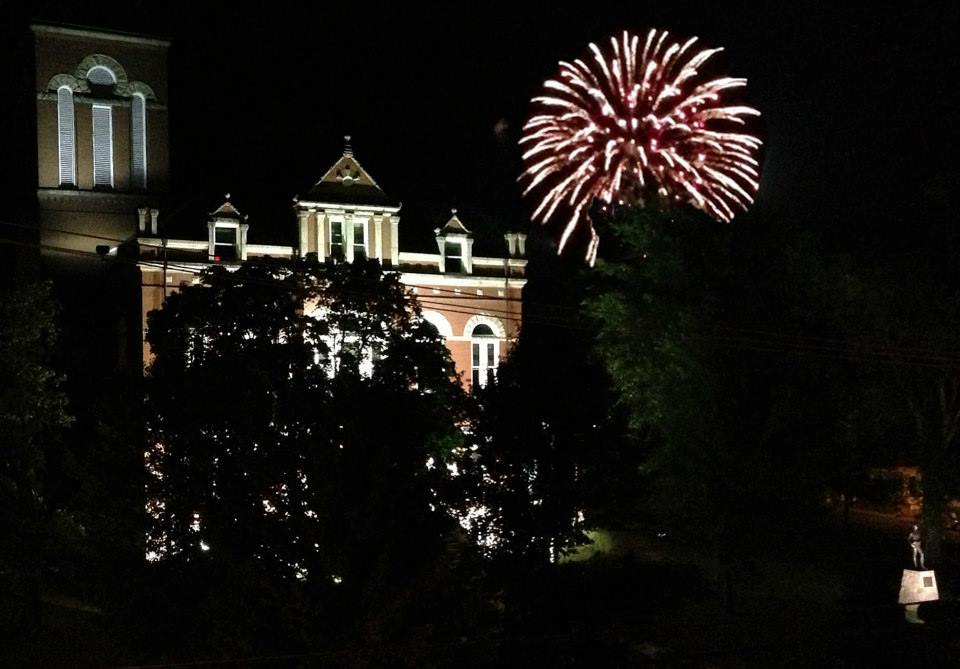 Colorful fireworks going off behind the lighted Fayette County Courthouse. The statue of Lafayette Flats is in the foreground. 