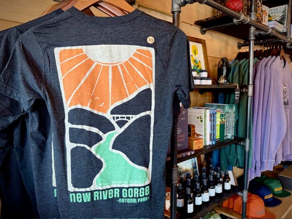 A dark t-shirt with a New River Gorge design in the foreground with other souvenirs like books, hats and art in the background.
