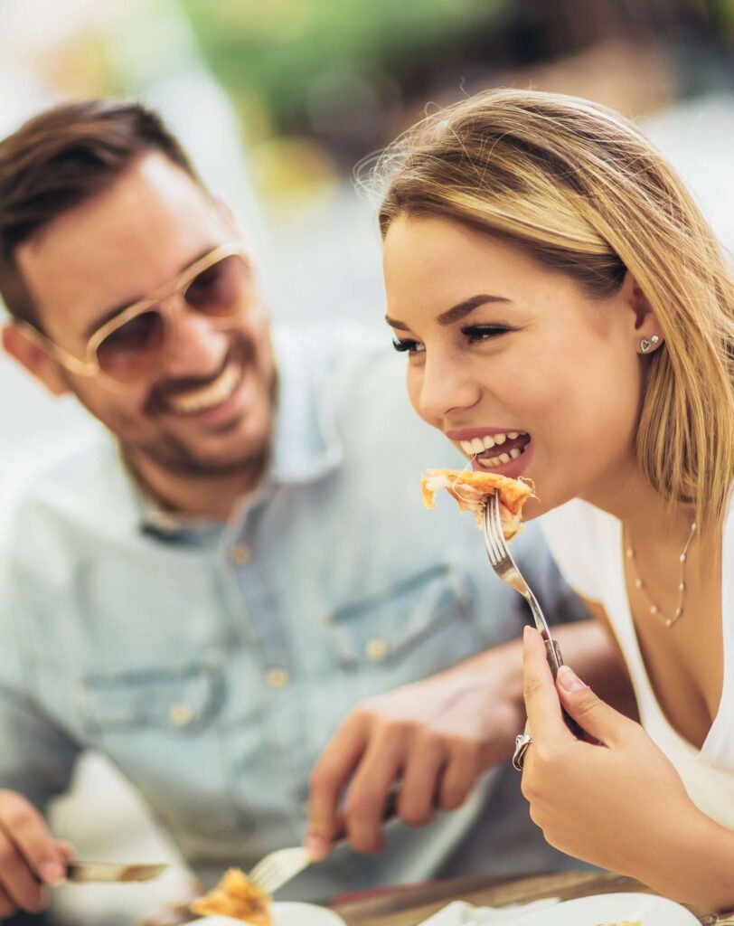 Woman enjoying a bite of pizza while her husband admires her on their anniversary getaway. 