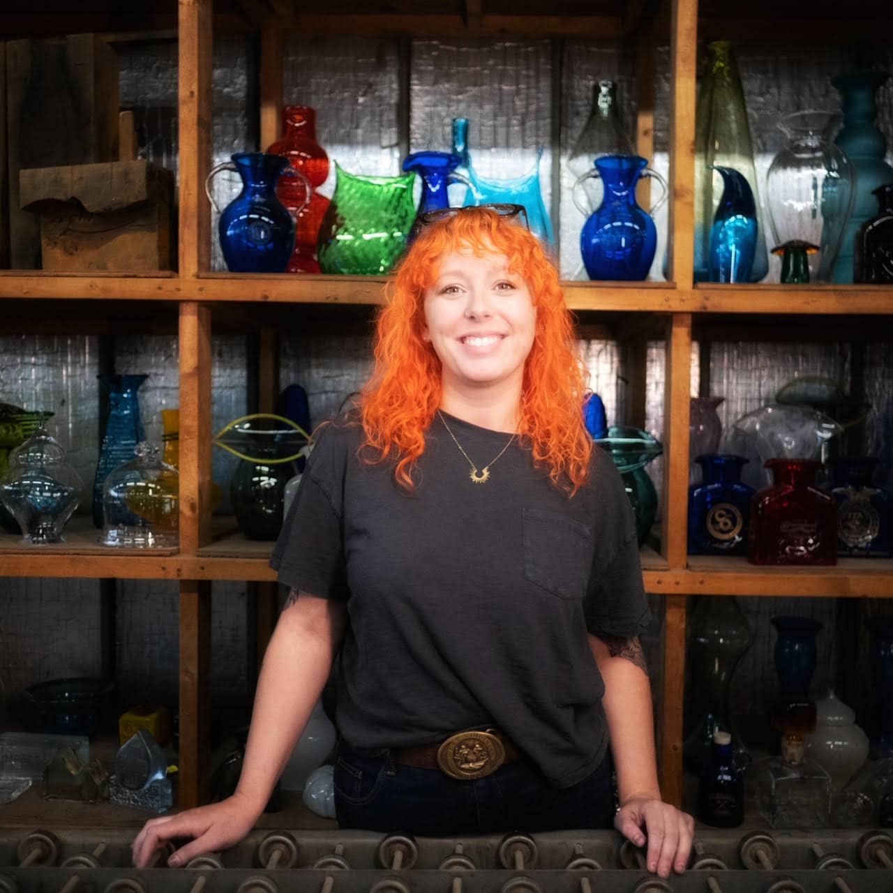 A smiling Odana Chaney (the 2024 New River Gorge Creative-in-Residence) with bright orange hair stands in front of shelf full of Blenko glass.