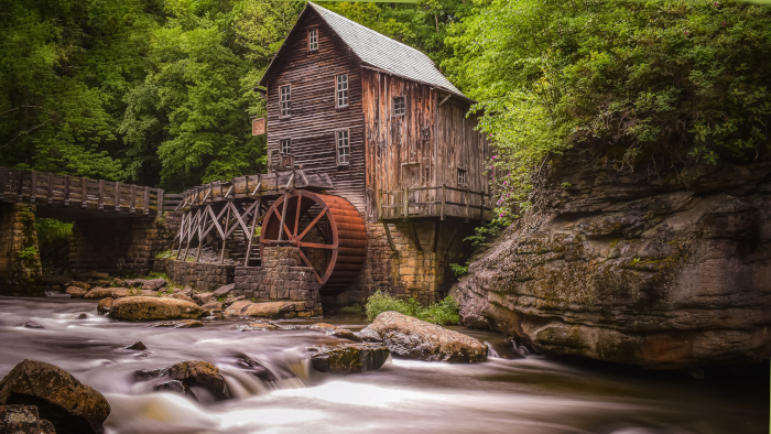 The Babcock Grist Mill with rushing water in the forground and green foliage in the background. 