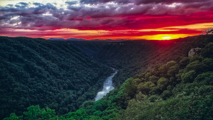 The vibrant colors of a cloudy sunset - purple, blue, violet, red, orange and yellow - above green mountains of the New River Gorge. 
