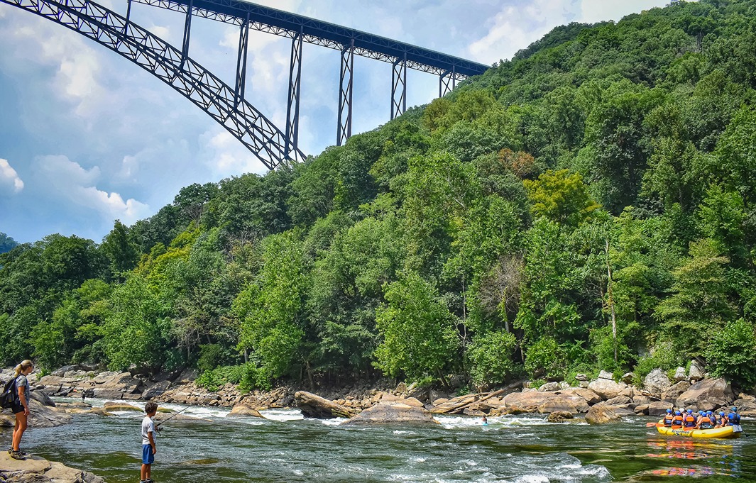 A young boy fishing on the side of the New River as a boat of whitewater rafters approach the rocky rapids of Fayette Station under the New River Gorge Bridge.
