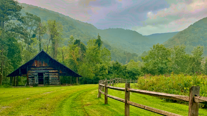 An wood fence leading to an old barn with lush green trees, fields and moutains all around. The Richmond-Hamilton Farm near Hinton. 