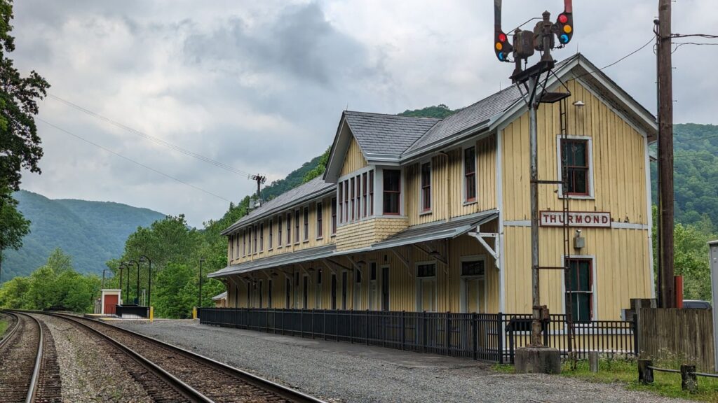 The yellow, two-story historic train depot in the forground with traintracks running through the photo into the green mountains in the distance. " The "Thurmond" sign is on the side of the building. 
