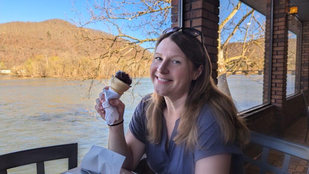 A girl (Amy) smiling while eating a dipped ice cream cone in front of Hinton Dairy Queen's windows which showcase the New River up close. 