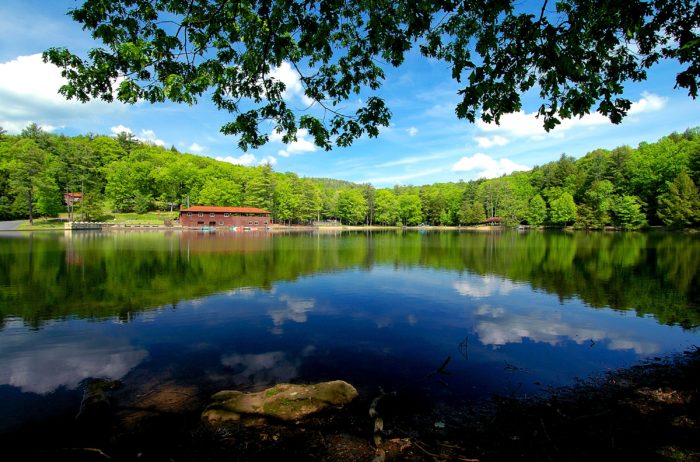 A shiny blue lake with clouds reflected in the water. The lush green trees of Little Beaver State Park surround the lake. 