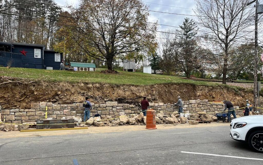 A very long and tall stone retaining wall being reconstructed by a group of five men. The photo was taken from inside Secret Sandwich Society. 