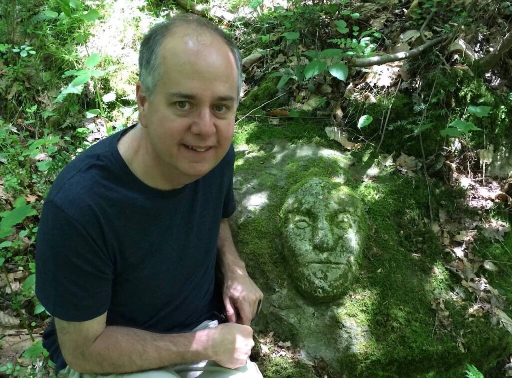 A man (Shawn) kneeling beside a large rock on the ground with a very distinct face carved into it. 
