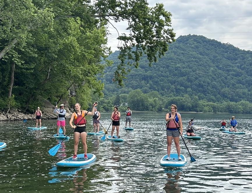 Numerous people standing and sitting on standup paddleboards in the middle of the New River with green leaves hanging overhead from the riverbank. 