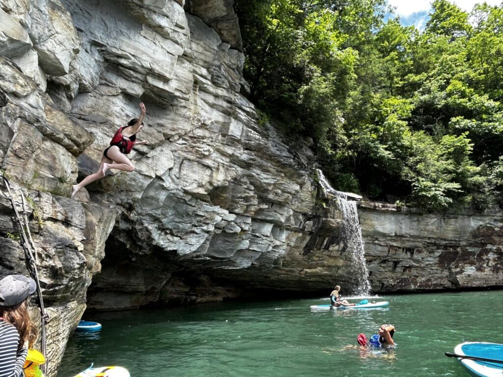 The clear water of Summersville Lake surrounded by white cliffs and green trees. A waterfall is in the center with people swimming and paddle boarding around it. 