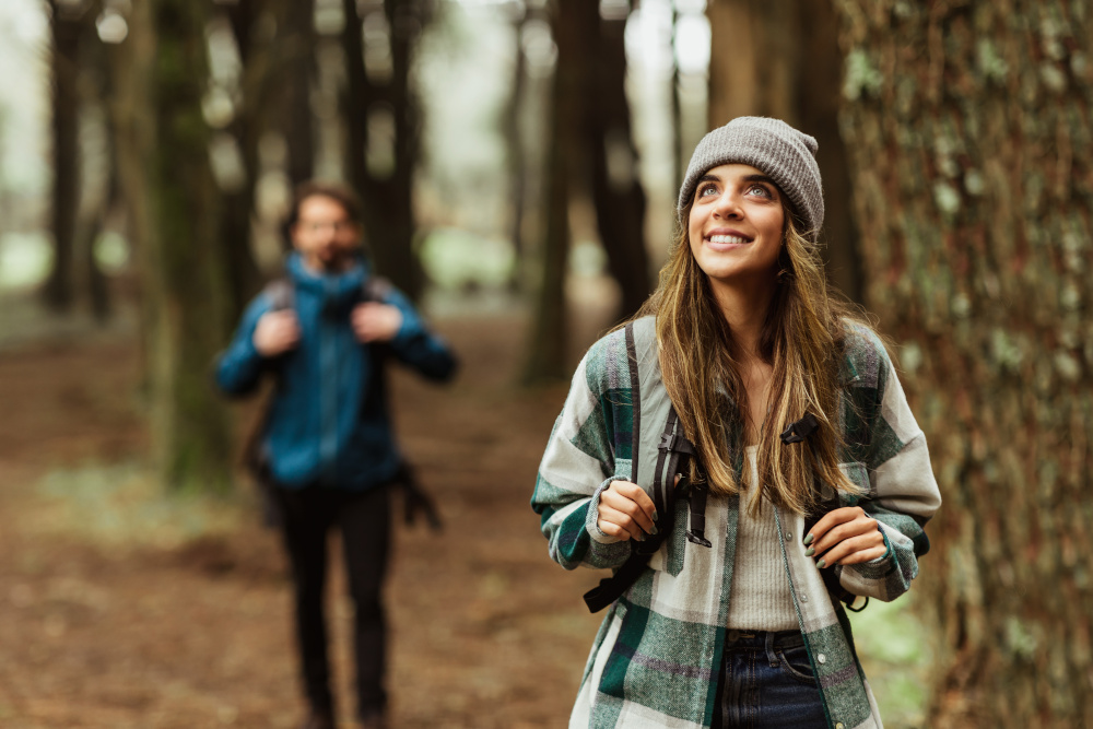 Smiling young woman in a flannel hoodie hiking in a fall forest with her boyfriend close behind.